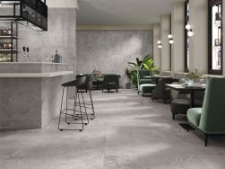 Restauracja w stylu industrialnym wyłożona szarymi płytkami imitującymi kamień Dome Ash z barem z dwoma wysokimi taboretami oraz stolikami z zielonymi fotelami