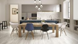 Jasna kuchnia z podłogą wyłożoną płytkami drewnopodobnymi z kolekcji Millelegni White Toulipier, z długim drewnianym stołem z kolorowymi krzesłami, niebieskimi i szarymi meblami oraz lampą wiszącą