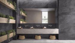 Ciemna łazienka wyłożona płytkami imitującymi kamień z kolekcji Nordika z wiszącą półką z trzema umywalkami i bateriami podtynkowymi, długim lustrem, trzema koszami wiklinowymi i półkami z roślinami
