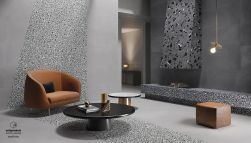 Nowoczesne pomieszczenie ze ścianą i podłogą wyłożoną jasnoszarymi płytkami lastryko z kolekcji Medley Grey, z kominkiem, brązowym fotelem i pufą oraz dwoma okrągłymi stolikami