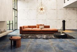 Salon, na środku duża kanapa w pomarańczowym kolorze, na podłodze dywan i czarny okrągły puf, dekoracyjna lampa, na podłoga wyłożona niebieskimi płytkami, na ścianie Flamed Ice Natural Rect. 49,75x99,55 płytki imitujące metal
