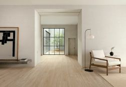 Przestronne, minimalistyczne pomieszczenie z jasną podłogą wyłożoną płytkami drewnopodobnymi Granier Maple, z fotelem i lampą stojącą oraz wiszącą półką z obrazem