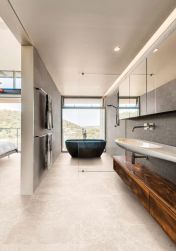 Podłużna łazienka z podłogą wyłożoną jasnymi płytkami imitującymi kamień Groove Hot White, z ciemną wanną wolnostojącą, kabiną prysznicową, umywalką wiszącą z drewnianą szafką i długim lustrem z szafką