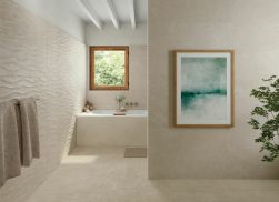 Beżowa łazienka ze ścianą wyłożoną dekoracyjnymi płytkami Ground Bone Decor z wanną pod oknem, ręcznikami na ścianie, obrazem i kwiatem
