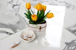 Zbliżenie na cztery płytki w połysku imitujące marmur białe z ciemnymi smugami Crash Marble Bianco 60x60 z ozdobnym żółtym kwiatkiem oraz cukierkami Carrea