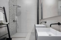 Łazienka, dwie umywalki z czarnymi podtynkowymi bateriami, na przeciwko wieszak na ręczniki w kolorze czarnym, prysznic, ścianka pod prysznicem wyłożona płytkami dekoracyjnymi przypominającymi drobną cegiełkę, ściany w łazience wyłożone szarymi płytkami L