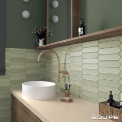 Fragment łazienki ze ścianą do połowy wyłożoną zielonymi cegiełkami z kolekcji Lanse i do połowy pomalowaną farbą, z drewnianą szafką z białą umywalką nablatową i baterią podtynkową oraz półką wiszącą z kosmetykami i kwiatami