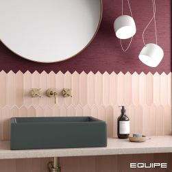 Widok na ścianę w łazience wyłożoną do połowy różowymi cegiełkami z kolekcji Lanse i pomalowaną fioletową farbą, z półką wiszącą z ciemną umywalką nablatową z baterią podtynkową, okrągłym lustrem i dwiema lampami wiszącymi