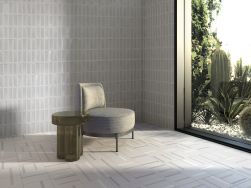 Pokój utrzymany w szarości ze ścianą wyłożoną szarymi płytkami trójwymiarowymi Levels Grey z szarym fotelem, okrągłym stolikiem i dużym oknem z widokiem na rośliny