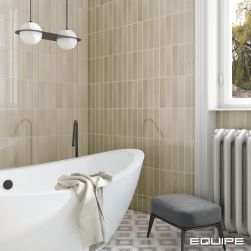Fragment łazienki ze ścianami wyłożonymi beżowymi cegiełkami z kolekcji Limit z podłogą wyłożoną jasnymi płytkami patchworkowymi, z białą wanną wolnostojącą z ręcznikiem, szarą pufą i lampą wiszącą