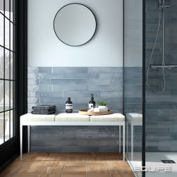 Łazienka z drewnopodobną podłogą i niebieską ścianą wyłożoną cegiełkami z kolekcji Manacor, z kabiną prysznicową, jasnym siedziskiem z kosmetykami oraz okrągłym lustrem