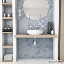 Widok na ścianę w łazience wyłożoną błękitnymi cegiełkami z kolekcji Manacor, z drewnianą półką wiszącą z białą umywalką, okrągłym lustrem i wąską szafką z boku z półkami