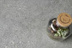 Szara betonowa płytka lastryko Manhattan Grey, a na niej szklany słoik z roślinami