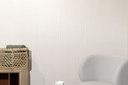 Zbliżenie na ścianę wyłożoną białymi, dekoracyjnymi płytkami ze żłobieniami Mare White Mix, z szarym fotelem i szafką z wiklinowym koszem z kocem