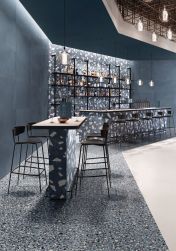 Restauracja ze ścianą wyłożoną ciemnymi, niebieskimi płytkami lastryko z kolekcji Medley Blue Minimal, z długim stołem, barem, czarnymi krzesłami i lampami wiszącymi