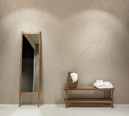 Ściana wyłożona wielkoformatowymi beżowymi płytkami imitującymi kamień Nature Beige ze stojącym podłużnym lustrem i drewnianą ławą ze szlafrokiem i wiklinowym dzbanem z ręcznikiem