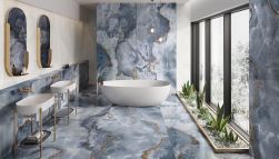Elegancka łazienka wyłożona niebieskimi płytkami imitującymi onyks z kolekcji Tele di Marmo z białą wanną wolnostojącą, dwiema umywalkami stojącymi, dwoma owalnymi lustrami oraz roślinnością pod oknami