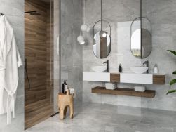 Łazienka wyłożona szarymi płytkami imitującymi kamień z kolekcji Wells z kabiną prysznicową, wiszącą półką z dwiema umywalkami nablatowymi, dwoma owalnymi lustrami, lampą wiszącą i białym szlafrokiem