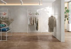 Sklep odzieżowy ze ścianami wyłożonymi jasnoszarymi płytkami imitującymi beton Koncept Nacar ze stołami i wieszakami z ubraniami