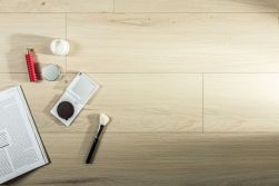 Widok z g&oacute;ry na podłogę z jasnych, dębowych paneli o subtelnym usłojeniu. Po lewej stronie ułożono kosmetyki do makijażu: paletkę z lusterkiem, pędzel, krem, czerwone perfumy oraz otwarty magazyn z tekstem.