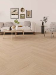 Jasny salon z podłogą ułożoną w jodełkę klasyczną o ciepłym odcieniu drewna. W tle stoi beżowa sofa z poduszkami, przed nią okrągły stolik kawowy. Na ścianie wisi galeria obraz&oacute;w, a po prawej widać fotel z puszystym, szarym futrem.
