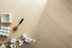 Zbliżenie z g&oacute;ry na podłogę z jasnych paneli ułożonych w klasyczną jodełkę. W lewym dolnym rogu kompozycji leżą dekoracje: gałązka bawełny z białymi puszystymi kwiatami, pędzel do makijażu oraz dwie białe, bąbelkowe świece strukturalne.
