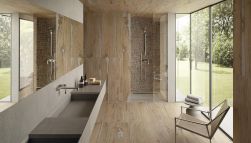 Rustykalna łazienka wyłożona płytkami drewnopodobnymi z kolekcji Alter Noce, z kabiną prysznicową, wiszącą półką z umywalką, długim lustrem, krzesłem i drzwiami prowadzącymi do ogrodu
