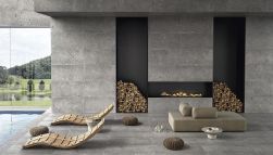 Taras z basenem wyłożony szarymi płytkami imitującymi kamień z kolekcji Ego Grigio Scuro z leżakami, kanapą i kominkiem w ścianie