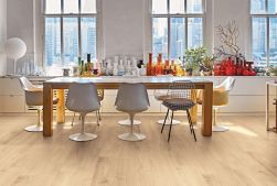 Jadalnia z podłogą wyłożoną beżowymi płytkami imitującymi drewno Provoak Rovere Puro, z długim stołem jadalnianym, krzesłami, trzema oknami i dużą ilością kolorowych wazonów na parapecie