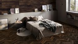 Ciemna sypialnia wyłożona płytkami drewnopodobnymi Provoak Nero Bruciato, z podwójnym łóżkiem, okrągłym stolikiem, oknem, obrazami i książkami
