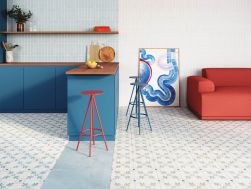 Kuchnia połączona z salonem ze ścianą wyłożoną niebieskimi cegiełkami Rabat Blue z niebieskimi meblami z drewnianymi blatami, wiszącą półką, czerwonym i dwoma niebieskimi taboretami, obrazem i czerwoną kanapą