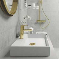 Biała umywalka nablatowa Deante Correo ze złotym korkiem i złotą baterią stojącą, na ścianie okrągłe lustro w złotej ramie, a obok kabina prysznicowa