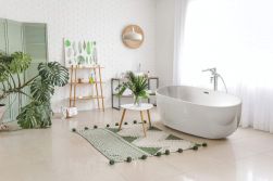 Przytulna łazienka z białą wanną wolnostojącą i baterią wolnostojącą w chromie Deante Alpinia, z dywanikiem, szafkami, i kwiatami