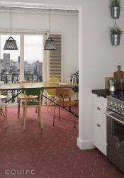 Kuchnia z czerwoną podłogą wyłożoną płytkami z kolekcji Rhombus z białymi meblami z czarnymi blatami, długim stołem jadalnianym z krzesłami, lampami wiszącymi i balkonem z widokiem na miasto