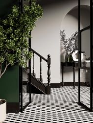 Elegancki korytarz z podłogą wyłożoną biało-czarną mozaiką Lordy Rosell Carrara z czarnymi schodami i czarnym sekretarzykiem z kwiatami i ozdobami, dużą rośliną w donicy oraz czarnymi, szklanymi drzwiami