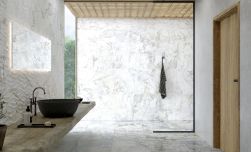 Łazienka z podłogą wyłożoną jasnymi płytkami imitującymi marmur Supreme White NT z marmurowymi ścianami, kabiną prysznicową walk-in, drewnianą półką z ciemną umywalką nablatową, lustrem i drzwiami