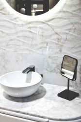 Zbliżenie na ścianę w łazience wyłożoną płytkami strukturalnymi imitującymi marmur Supreme White Decor z okrągłym lustrem, białą umywalką nablatową na marmurowym blacie szafki i małym lusterkiem