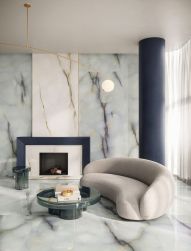 Salon wyłożony błękitnymi płytkami imitującymi kamień onyks z kolekcji Tele di Marmo Pure Onyx z kominkiem, szarą kanapą, okrągłym stolikiem i lampą wiszącą