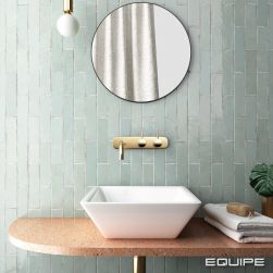 Ściana w łazience wyłożona miętowymi cegiełkami z kolekcji Tribeca z kolorowym, wiszącym blatem z białą, geometryczną umywalką nablatową, złotą baterią podtynkową, okrągłym lustrem i ręcznikami