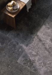 Widok na podłogę w łazience wyłożoną antracytowymi płytkami imitującymi kamień z kolekcji Unique Bleu Ancienne Anthracite, z drewnianą ławą z ręcznikiem i przyborami do mycia