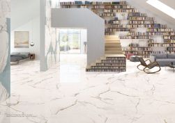 Duże, jasne pomieszczenie wyłożone płytkami imitującymi marmur z kolekcji Unique Marble Marmo Paonazzetto, ze schodami, regałami z książkami, szarą kanapą i fotelem