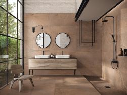 Łazienka wyłożona jasnobrązowymi płytkami imitującymi metal lappato Britannic Copper z prysznicem, szafką z dwiema umywalkami nablatowymi, dwoma okrągłymi lustrami i fotelem
