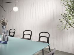 Pokój ze ścianą wyłożoną płytkami z kolekcji Esencia Material z błękitnym stołem, trzema czarnymi krzesłami i lampą wiszącą