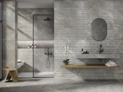 Łazienka wyłożona szarymi płytkami imitującymi kamień z reliefem Caesar Grey z kabiną prysznicową, umywalką ścienną i owalnym lustrem