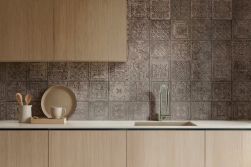 Nowoczesna kuchnia z szafkami w kolorze jasnego dębu i białym blatem. Ścianę zdobią brązowe, wzorzyste kafle typu patchwork. Na blacie widoczny jest ceramiczny kubek, talerz oraz drewniane akcesoria. Całość dopełnia minimalistyczny zlew i bateria.