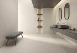 Nowoczesny salon kąpielowy w jasnych płytach. Po lewej stoi szara, tapicerowana ławka. Po prawej długi, betonowy blat z dwiema grafitowymi umywalkami, nowoczesną armaturą i owalnymi lustrami. W głębi widać podświetlaną wnękę z p&oacute;łkami.
