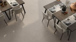 Widok z g&oacute;ry na jasne, matowe płyty podłogowe w jadalni lub restauracji. Po obu stronach kadru stoją ciemne drewniane stoły z kompletną zastawą i bieżnikami, przy kt&oacute;rych ustawiono eleganckie, tapicerowane krzesła w odcieniu złamanej bieli.