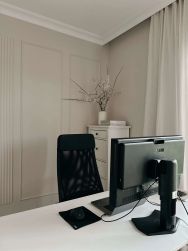 Nowoczesne biuro domowe w jasnych, beżowych barwach. Na pierwszym planie białe biurko z czarnym monitorem NEC i fotelem. W tle elegancka ściana z sztukaterią oraz biała komoda z wazonem z gałązkami.