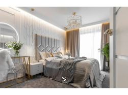 Luksusowa sypialnia w stylu glamour. Centralne miejsce zajmuje duże ł&oacute;żko z szarym, pikowanym zagł&oacute;wkiem. Obok toaletka i kryształowy żyrandol. Ciepłe światło LED oświetla geometryczną tapetę.