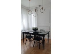 Elegancka jadalnia z czarnym stołem i krzesłami patyczakami. Na ścianie klasyczna biała sztukateria. Nad stołem wiszą minimalistyczne, okrągłe lampy, a w tle widać okno z białą firaną.